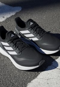 Chaussures de sport noires avec des accents rayés blancs, tige texturée, semelles épaisses et design léger, positionnées sur une surface en asphalte.