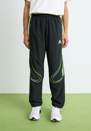 Mand iført sorte Adidas træningsbukser med neongrøn piping og hvide sneakers, stående på grøn gulv imod hvid væg.