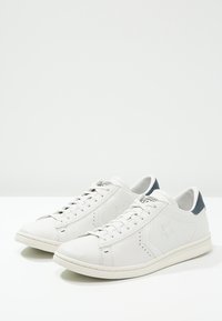 Baskets en cuir blanc avec un accent texturé bleu marine sur le talon, des détails perforés et une semelle en caoutchouc plate. Design simple avec des lacets.