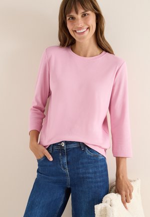 Top de manga larga de color rosa claro con textura acanalada y escote redondo, combinado con unos jeans de color azul oscuro y sosteniendo un bolso crema. Diseño sencillo y textura suave.