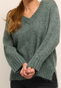 Un pull en tricot vert doux avec un col en V, des poignets côtelés et des fentes latérales. Le tissu semble texturé et lâchement tricoté pour un ajustement décontracté.