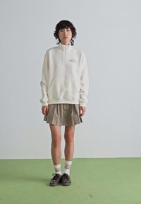 Une personne avec des cheveux courts et bouclés porte un sweat-shirt blanc à fermeture éclair, une jupe plissée beige, des chaussettes rayées et des baskets noires, debout sur un sol vert.