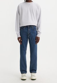 Man som bär en ljusgrå sweatshirt, blå raka jeans och vita sneakers, stående mot en vit bakgrund.