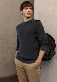 Pull gris foncé en maille avec encolure ronde et poignets côtelés, associé à un pantalon beige clair. Un sac à dos noir est porté sur une épaule.