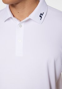 Vit pikétröja med mjuk textur, med en knappslå och en marinblå logotyp på kragen. Designen är figursydd och elegant.