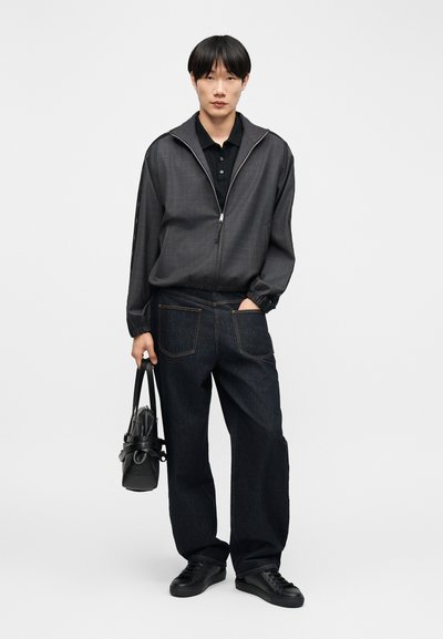 Νεαρός άνδρας που φοράει μαύρο μπουφάν με φερμουάρ, μαύρο πόλο, φαρδιά σκούρα τζιν, μαύρα αθλητικά παπούτσια, κρατώντας μια μαύρη τσάντα στο ένα χέρι.