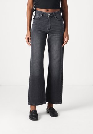 Personne portant un jean noir taille haute à jambes larges, associé à des mocassins noirs, debout devant un fond blanc uni.