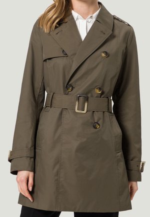 Femme portant un trench-coat vert olive croisé avec ceinture et poignets boutonnés sur une chemise blanche à col.