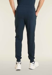 Pantalones de chándal azul marino con puños acanalados, cinturilla elástica y detalle de bolsillo trasero, combinados con zapatillas deportivas blancas en un entorno minimalista.