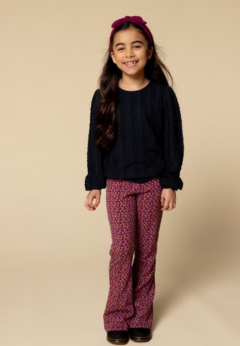 Zwarte gebreide trui met een textuurpatroon, gecombineerd met uitgezakte paarse broeken met een kleurrijke abstracte print. Donkere schoenen en een roze haarband.