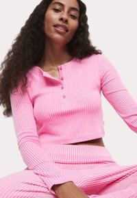 Roze geribbelde crop top met lange mouwen, voorzien van een ronde halslijn en een viervoudige knopenlijst. De stof heeft een gestructureerde uitstraling en gevoel.
