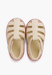 Beige rubber sandals with a closed heel and open toe design. Features cut-out patterns and a pink textured sole. Lightweight and flexible.