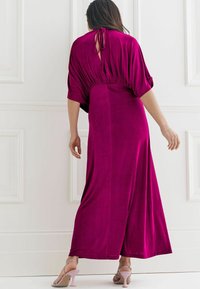 Robe maxi fuchsia avec manches courtes drapées, taille cintrée et détail œillet au dos. Texture lisse et douce avec une silhouette fluide.