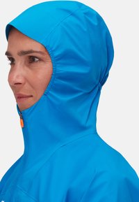 Donna che indossa un giaccone a zip di un luminoso blu, chiuso fino al mento, con un primo piano del profilo laterale su uno sfondo bianco.