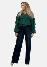 Zielona, falbaniasta bluzka z guzikami i długimi rękawami, w zestawie z jeansami o wysokim stanie w ciemnym odcieniu. Modelka stoi na neutralnym tle.