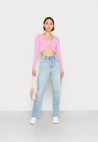 Top cropped de manga longa em rosa com decote em V e detalhe torcido, combinado com jeans de cintura alta em azul claro e uma mini mala floral.