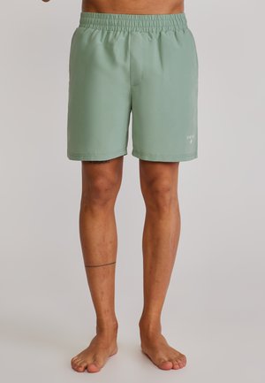 Mann trägt hellgrüne Shorts mit elastischem Bund, steht barfuß vor einem einfarbigen Hintergrund.