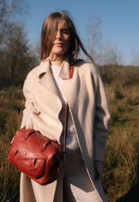 Red leather handbag with rounded shape, zip closure, dual top handles, and adjustable shoulder strap. Worn against a light coat.