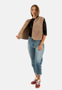 Gilet in pile beige con una fodera liscia e bottoni, indossato sopra una camicia nera a fantasia. Il modello indossa anche jeans azzurri arrotolati e sneakers rosse.