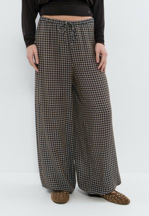 Persona con camiseta negra de manga larga, pantalones de cuadros de pierna ancha con cordón y zapatos marrones sin cordones con detalles de ojales plateados, de pie contra un fondo blanco.