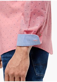Camicia a pois rossi e blu con polsini blu chiaro, caratterizzata da dettagli sui bottoni e tessuto testurizzato. Mano appoggiata su jeans di denim.