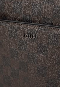 Brunt rutete lærmateriale med en teksturert overflate. Har en liten "JOOP!"-logo i sølv og subtile mønsterdetaljer.