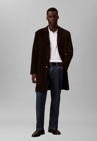 Fekete hosszú kabát fehér pólóinggel, kék farmernadrággal és fekete bőrcipővel párosítva. Klasszikus gallérral és gombos lezárással rendelkezik.