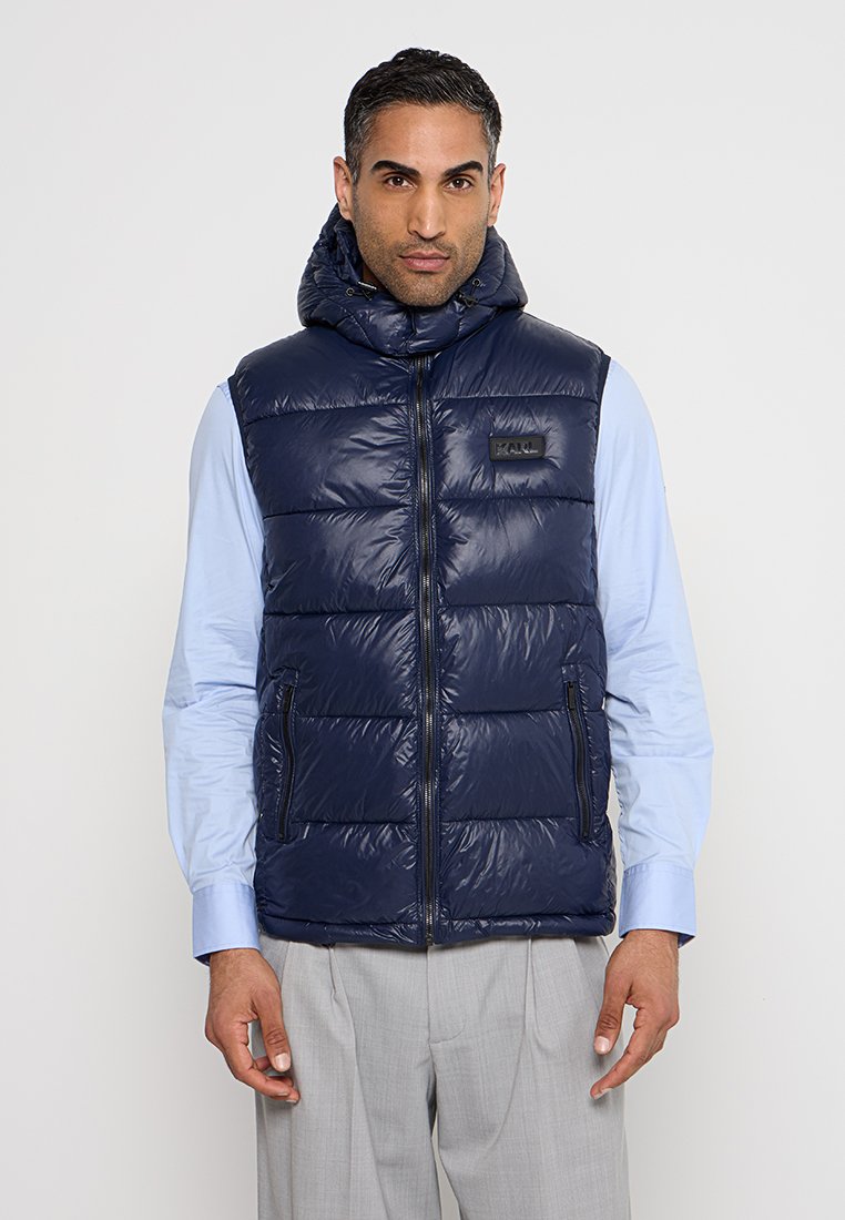 Gilet matelassé bleu marine avec capuche, texture matelassée et fermeture éclair à l'avant. Comprend des poches zippées sur les côtés et un petit logo de la marque sur la poitrine.
