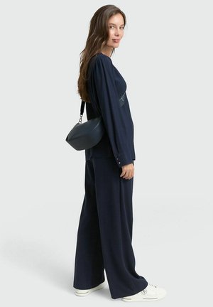 Marineblaues Langarmoberteil mit gefältelten Akzenten, kombiniert mit weiten Hosen. Accessoire umfasst eine runde schwarze Tasche und weiße Sneaker.