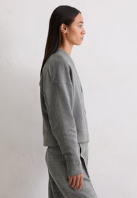 Cardigan court gris en tissu tricot doux, avec col rond, manches longues et fermetures à boutons, associé à un pantalon gris assorti.