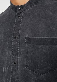 Camicia di denim nera con colletto rotondo, caratterizzata da una leggera texture, una chiusura frontale con bottoni e una tasca sul petto con un'etichetta logo.