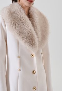 Donna che indossa un cappotto bianco con un ampio collare di pelliccia sintetica beige e bottoni dorati lucidi, mostrata dalle spalle alla vita.