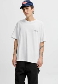 Balta īsām piedurknēm T-krekls ar apaļu kakla izgriezumu, kuram ir neliels sarkans logotips kreisajā pusē. Valkāts ar melniem biksēm un tumši zilā cepurē.