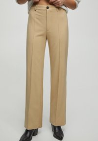 Pantalons larges beiges en tissu doux, avec fermeture à bouton sur le devant, des coutures verticales et une coupe ajustée.