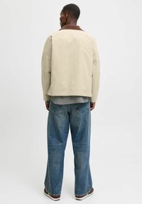 Beige jakke med brun krave, parret med løse blå denim jeans. Outfitten har en ternet skjorte, der er synlig ved kanten.