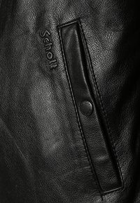 Surface en cuir noir texturé avec une poche verticale munie d'un bouton-pression et un logo « Schott NYC » embossé à proximité.