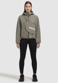 Padded olive green jacket with a zip closure and large flap pocket featuring "KHUJO" logo. Paired with black leggings and sneakers.