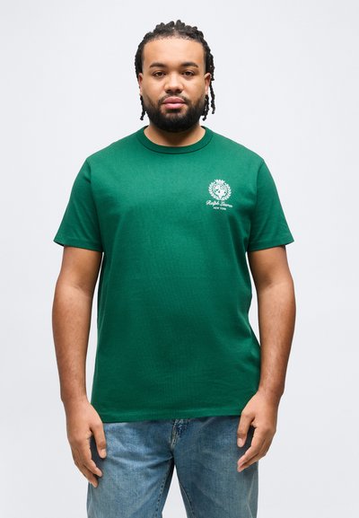 Άνδρας με πλεγμένα μαλλιά και γένια, φοράει πράσινο μπλουζάκι Ralph Lauren και μπλε τζιν, στέκεται μπροστά σε απλό ανοιχτό φόντο.