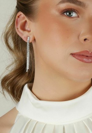 Silver rhinestone hängeörhängen med en smal, kaskad design. Bärs av en modell med en slät, neutralfärgad blus och texturerat hår.