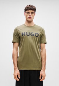 Olivovo zelené tričko s krátkym rukávom vyrobené z ľahkej bavlny, s čiernym logom "HUGO" na hrudi. Jednoduchý dizajn s okrúhlym výstrihom.