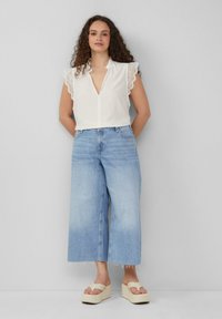 Chemisier blanc avec des manches en dentelle scallopée, associé à un jean large bleu clair et des sandales à plateforme beige avec une semelle épaisse.