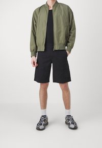 Giacca bomber verde oliva, pantaloni cargo neri, scarpe da ginnastica grigie con dettagli gialli e calzini bianchi. Abbigliamento casual e sportivo.
