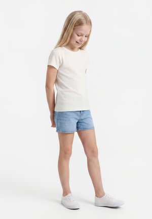 Young girl smiling, wearing a plain white short-sleeve shirt, blue denim shorts, and white sneakers, standing against a white background.