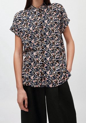 Žena nosící krátký rukáv s límečkem s vícebarevným květinovým vzorem a černé široké kalhoty, jedna ruka v kapse.