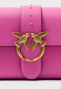 Rózsaszín bőr kézitáska, arany színű kör alakú fémrészlet két madár díszítéssel, tüskékkel dekorálva a fedélen a részletekért.