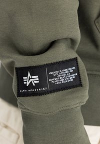 Olivno zeleno sweatshirt z črno oznako z belim besedilom, logotipom in šivi, z gladko in mehko teksturo.