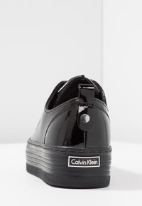 Black patent leather platform sneaker with pull tab and Calvin Klein logo on the back sole, shown from the rear angle.