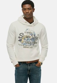 Sudadera blanca con un bolsillo canguro, que presenta un diseño gráfico con texto en azul, negro y amarillo. Combinada con jeans de mezclilla azules y un cinturón marrón.