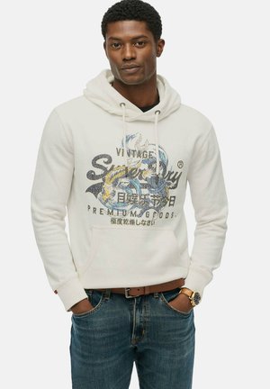 White hoodie with a kangaroo pocket, featuring a graphic design in blue, black, and yellow text. Paired with blue denim jeans and a brown belt.