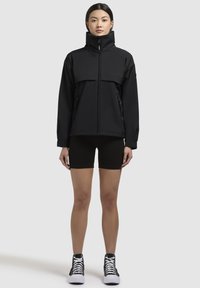 Giacca nera con colletto alto, chiusura zip e due tasche anteriori. Indossata sopra pantaloni corti neri e abbinata a scarpe sneakers in bianco e nero.